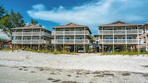 A beachfront hotel with multiple balconies, a sandy beach in the foreground, and clear skies. - Shore Haven Unit 5A by Distinctive Beach Rentals (Indian Shores)