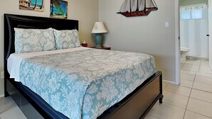A bedroom with a bed, a nightstand, a lamp, and two wall paintings. - Shore Haven Unit 5A by Distinctive Beach Rentals (Indian Shores)