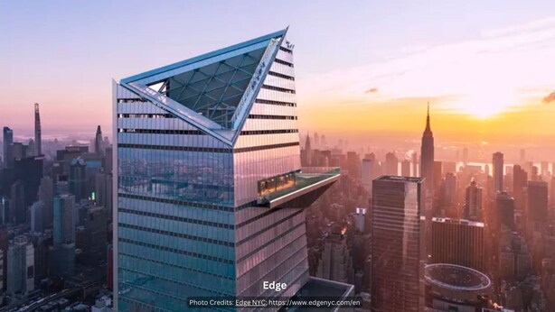 Een modern wolkenkrabber met een opvallend driehoekig dak en een glazen gevel, voor een stadsachtergrond tijdens zonsondergang.