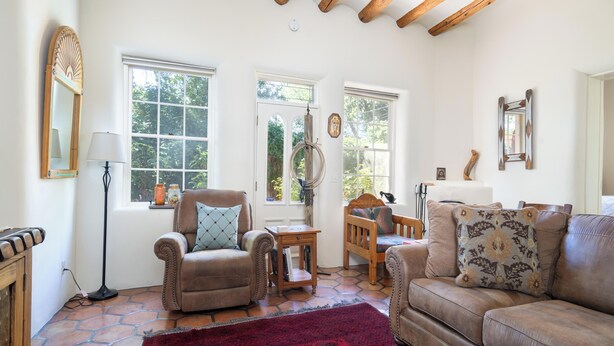 A cozy living room with a leather sofa, a wooden chair, a red rug, and large windows with views of greenery. - Siete Caballos by Kokopelli Property Management (Santa Fe)