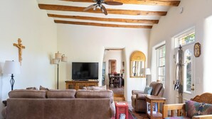 A living room with a large sofa, a wooden coffee table, and a television. There is a wooden ceiling with exposed beams and a cross on the wall. - Siete Caballos by Kokopelli Property Management (Santa Fe)