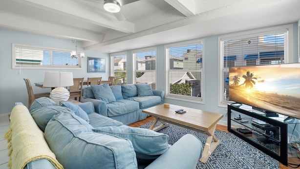 A living room with a sectional sofa, a coffee table, a TV on a stand, and large windows with blinds. - Sunset Beach House by Distinctive Beach Rentals (Treasure Island)