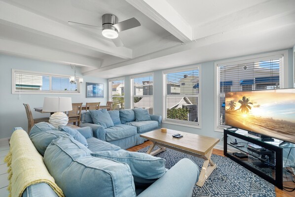 A living room with a sectional sofa, a coffee table, a TV on a stand, and large windows with blinds.