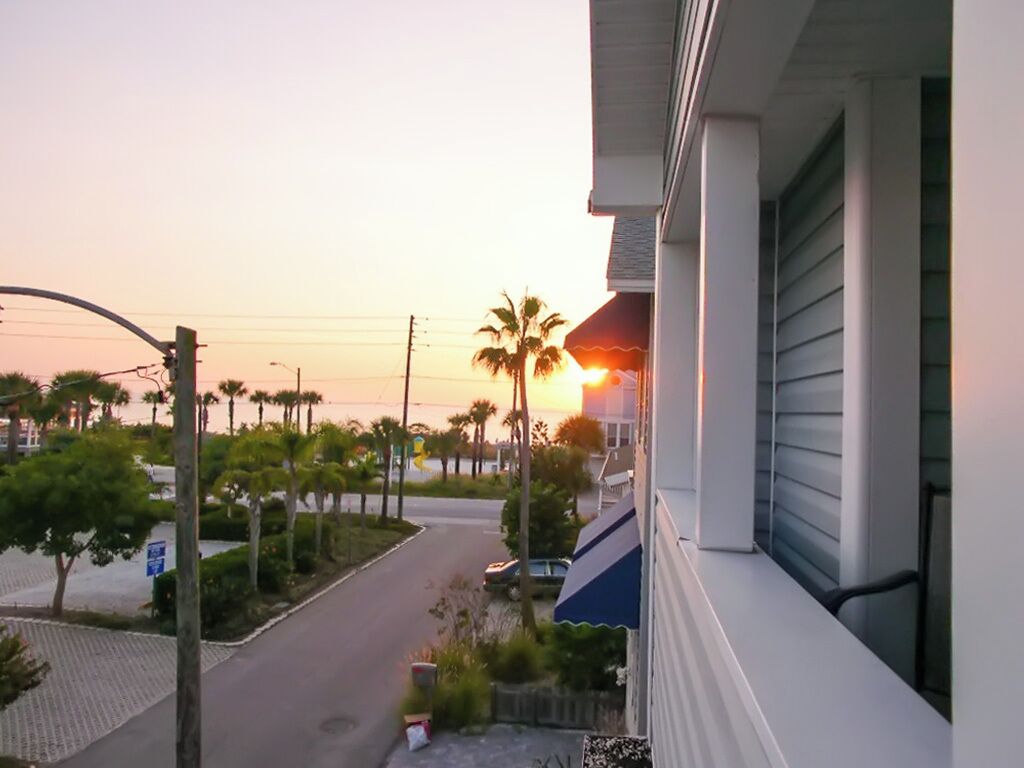 Een woonstraat met een huis aan de rechterkant, een palmboom en een zonsondergang op de achtergrond.