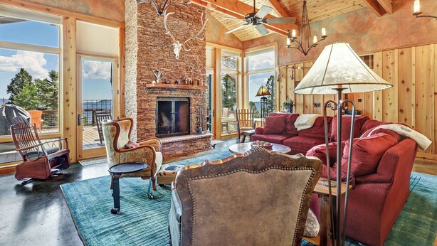 A spacious living room with a fireplace, wooden ceiling beams, and large windows offering a view of the outdoors.