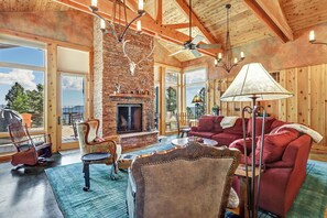 A spacious living room with a fireplace, wooden ceiling beams, and large windows offering a view of the outdoors. - Custom Luxury Cabin | Private Setting | Large Deck with Views | Jung's Bärenhaus (Angel Fire)