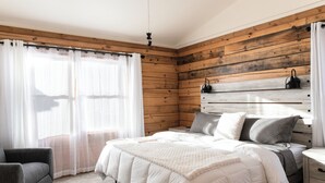 Une chambre avec un mur en bois, un lit avec une literie blanche, une table de chevet, une chaise et un ventilateur de plafond.
