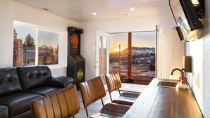 Un salon moderne avec un canapé en cuir noir, des fauteuils en cuir marron, un jeu de fléchettes et une vue sur un paysage enneigé par une porte vitrée.