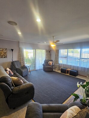 Ein modernes Wohnzimmer mit einem grauen Ecksofa, einem grauen Sessel und einem grauen Fernsehsessel. Es gibt einen Deckenventilator, eine Stehlampe und ein großes Fenster mit Vorhängen.