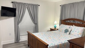 Un dormitorio con una cama de madera, cortinas grises, un televisor montado en la pared y una lámpara de noche.