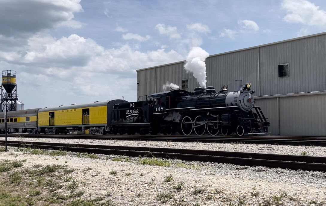 Una locomotora de vapor con 'U.S. Sugar' y '148' en el costado, arrastrando un tren de pasajeros amarillo.