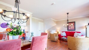 A dining area with a wooden table, pink chairs, and a chandelier. There is a red sofa and a wicker chair in the living area. - Haus Raanwai - Wattblick. Natur. Familie. - Rhr/3a (Sylt)