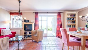 A living room with a red sofa, wicker armchair, and a glass coffee table. There's a TV on a wooden stand, a bookshelf, and a dining area with a table and chairs. - Haus Raanwai - Wattblick. Natur. Familie. - Rhr/3a (Sylt)