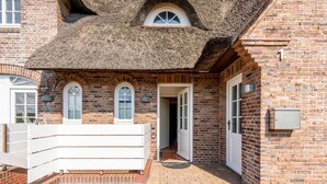 A brick building with a thatched roof, white doors, and a white balcony. - Haus Raanwai - Wattblick. Natur. Familie. - Rhr/3a (Sylt)