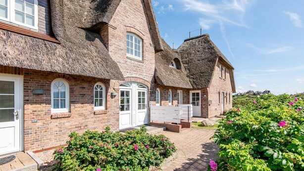 A row of thatched-roof houses with brick walls and white doors, surrounded by lush greenery and flowers. - Haus Raanwai - Wattblick. Natur. Familie. - Rhr/3a (Sylt)