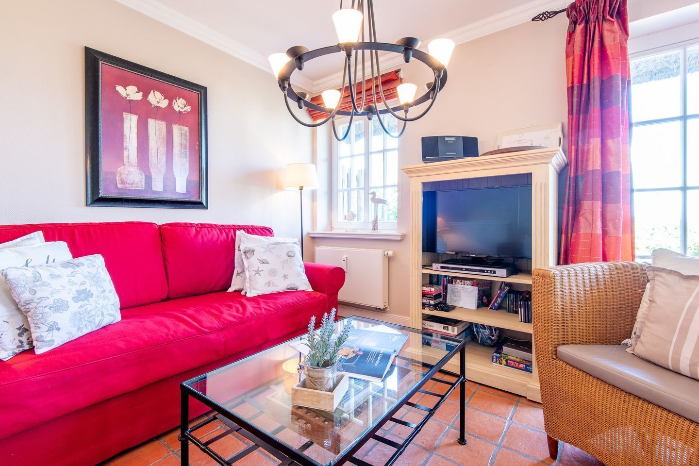 Una sala de estar con un sofá rojo, una mesa de centro de vidrio, un sillón de mimbre, una estantería y un televisor.