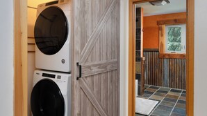Une buanderie équipée de deux machines à laver à chargement frontal superposées, d’une porte coulissante en bois et donnant sur une pièce avec une fenêtre et une rambarde en bois.