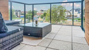 A modern balcony with a glass enclosure, wicker furniture, and a view of the cityscape.