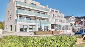 A multi-story hotel with a modern design, featuring a glass facade, multiple balconies, and a well-maintained outdoor area with hedges. - Haus Sommertraum, Appartement 5 "sonnensegel" - Haus Sommertraum (Scharbeutz)