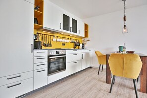 Uma cozinha moderna com armários brancos, forno embutido e painel de parede amarelo. Há uma área de jantar com uma cadeira amarela e uma mesa de madeira.