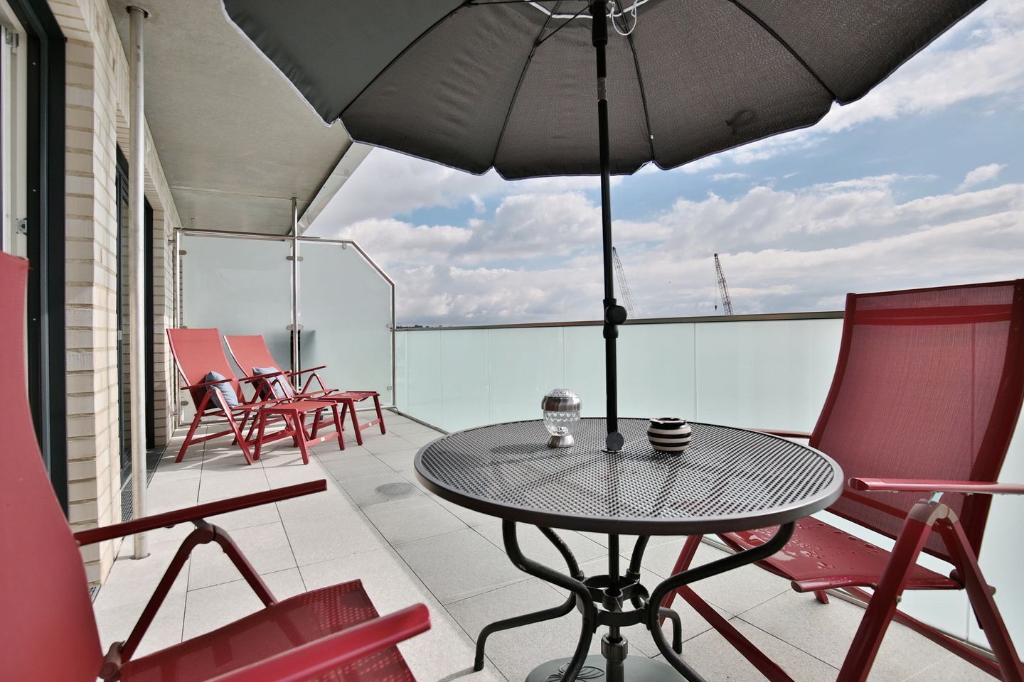 A rooftop terrace with a round table and red chairs, an umbrella, and a view of the city skyline.