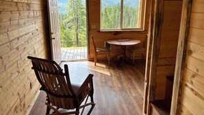 L’intérieur d’un chalet en bois comprend un coin repas, un fauteuil à bascule et une porte donnant sur un balcon avec vue sur la forêt.