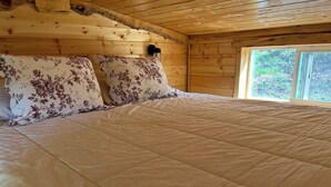 Une chambre dans une cabane en bois, avec un grand lit, des coussins à motifs floraux et une fenêtre donnant sur un paysage verdoyant.