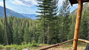 Une terrasse en bois avec une chaise noire, entourée d’une forêt et avec des montagnes en arrière-plan.