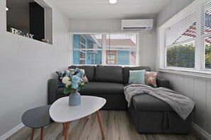 A modern living room with a grey sofa, a small white table, and a vase of flowers. There are windows with blinds and an air conditioning unit on the wall. - 70 Steps From The BEACH! W/ Grill & Hammock! Free parking!  (Clearwater)