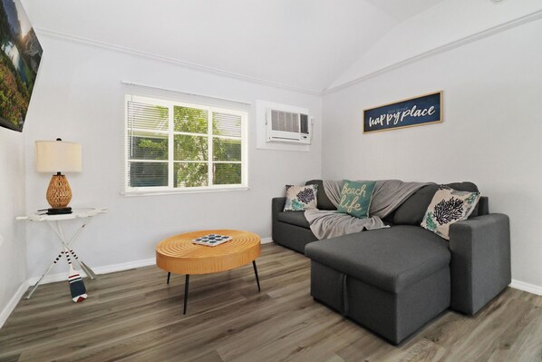 A modern living room with a gray sofa, a round wooden coffee table, a side table with a lamp, and a large window with blinds.