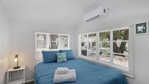 Un dormitorio con una cama, dos ventanas con persianas, una mesita de noche con lámpara y un aire acondicionado de pared.