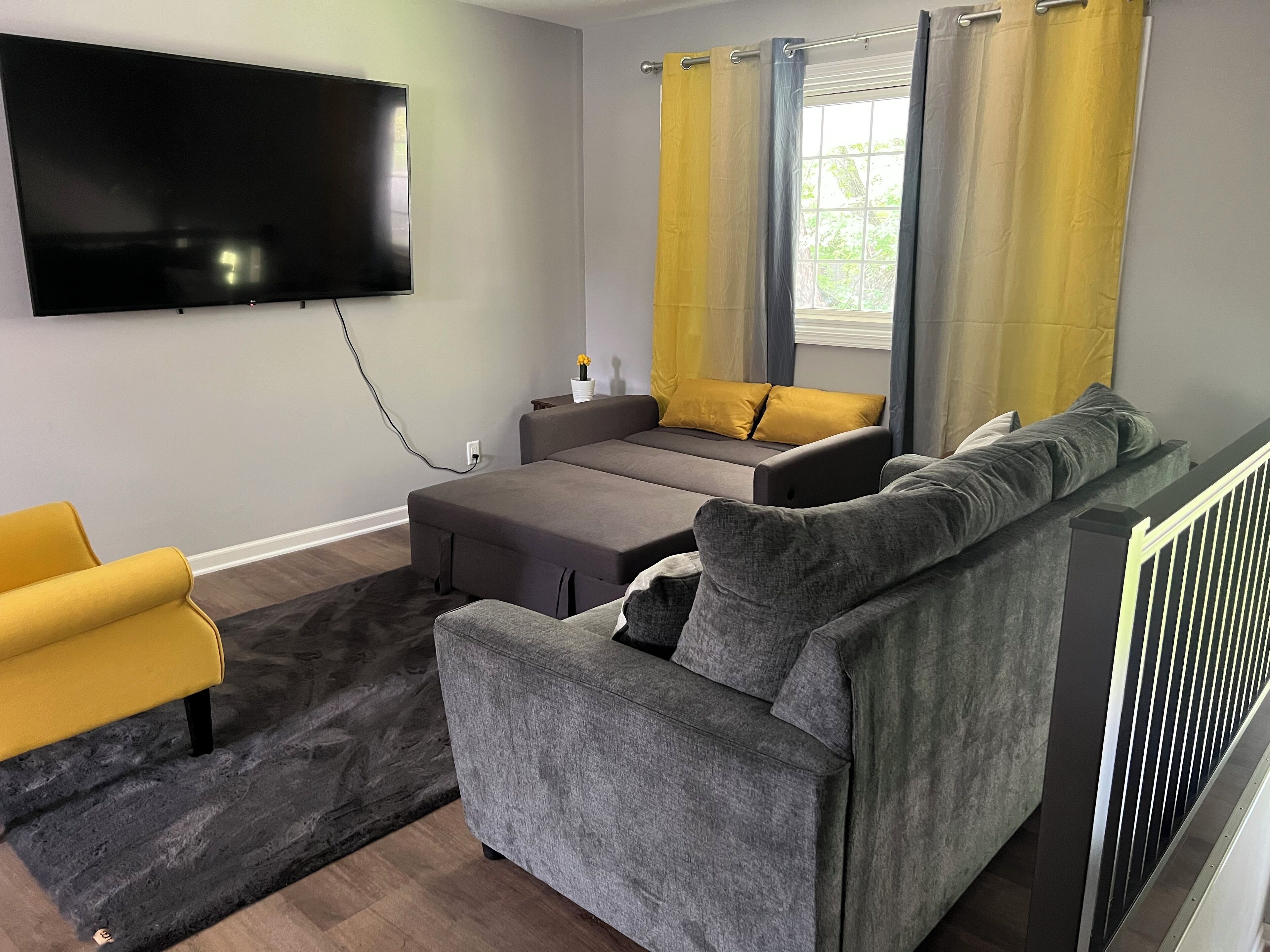 Een moderne woonkamer met een flatscreen televisie aan de muur, een grijze bank, een gele fauteuil en een klein bijzettafeltje met een bloemenvaas.