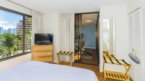 Quarto de hotel com cama, televisão sobre cômoda de madeira, varanda com vista para palmeiras e edifícios, e porta deslizante que dá para outro quarto.