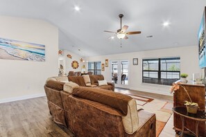 A spacious living room with a large brown sectional sofa, a ceiling fan, and a view through the glass doors to the outside.