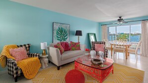 Een woonkamer met een blauwgroene wand, een geruite fauteuil, een bank met kleurrijke kussens, een glazen salontafel en uitzicht op zee door grote ramen.