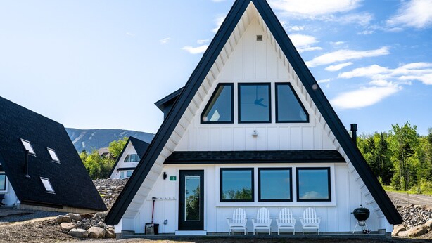 A white, triangular-roofed building with a black roof, two white chairs outside, and a black grill. - Saddleback - Parmachenee A-Frame #1 - SkiFrame (Sandy River Plantation)