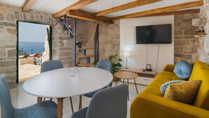 Un salon chaleureux avec un mur en pierre, des poutres en bois, une table ronde, un canapé jaune et un téléviseur fixé au mur.