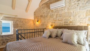 Une chambre avec un mur en pierre, un lit recouvert d’une couette à carreaux, une fenêtre donnant sur un beau paysage et un appareil de climatisation.