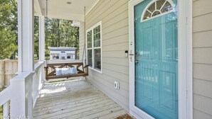 A porch with a swing, a teal door, and a view of a house in the background. - Only TWO MILES to the beach! Custom bungalow. Fenced-in yard! (Ocean Isle Beach)