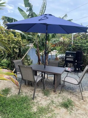 An outdoor patio with a table and chairs under a blue umbrella, surrounded by lush greenery and a barbecue grill. - Don Julio’s getaway home.<br>A nice place to relax next to mother’s nature <br> (luquillo)
