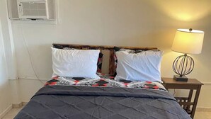 Una cama con colcha oscura, almohadas blancas y cubrecama estampado. Hay una mesita de noche con una lámpara y un aire acondicionado instalado en la pared.