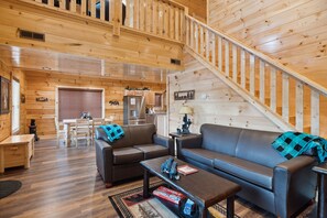 Cozy cabin interior with a high ceiling, sofa, coffee table, and dining table for two, plus a staircase and kitchen with modern appliances.