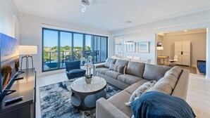 A modern living room with a large sofa, a coffee table, and a TV. There is a balcony with a view of the outdoors.