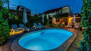 Piscina con sillones de descanso y sombrillas, rodeada de vegetación y un edificio con tejado rojo.