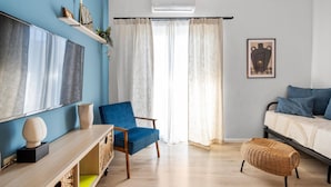 Una sala moderna con una pared azul de acento, un televisor de pantalla plana grande, un sillón azul, un reposapiés de mimbre y una cama con almohadas azules.