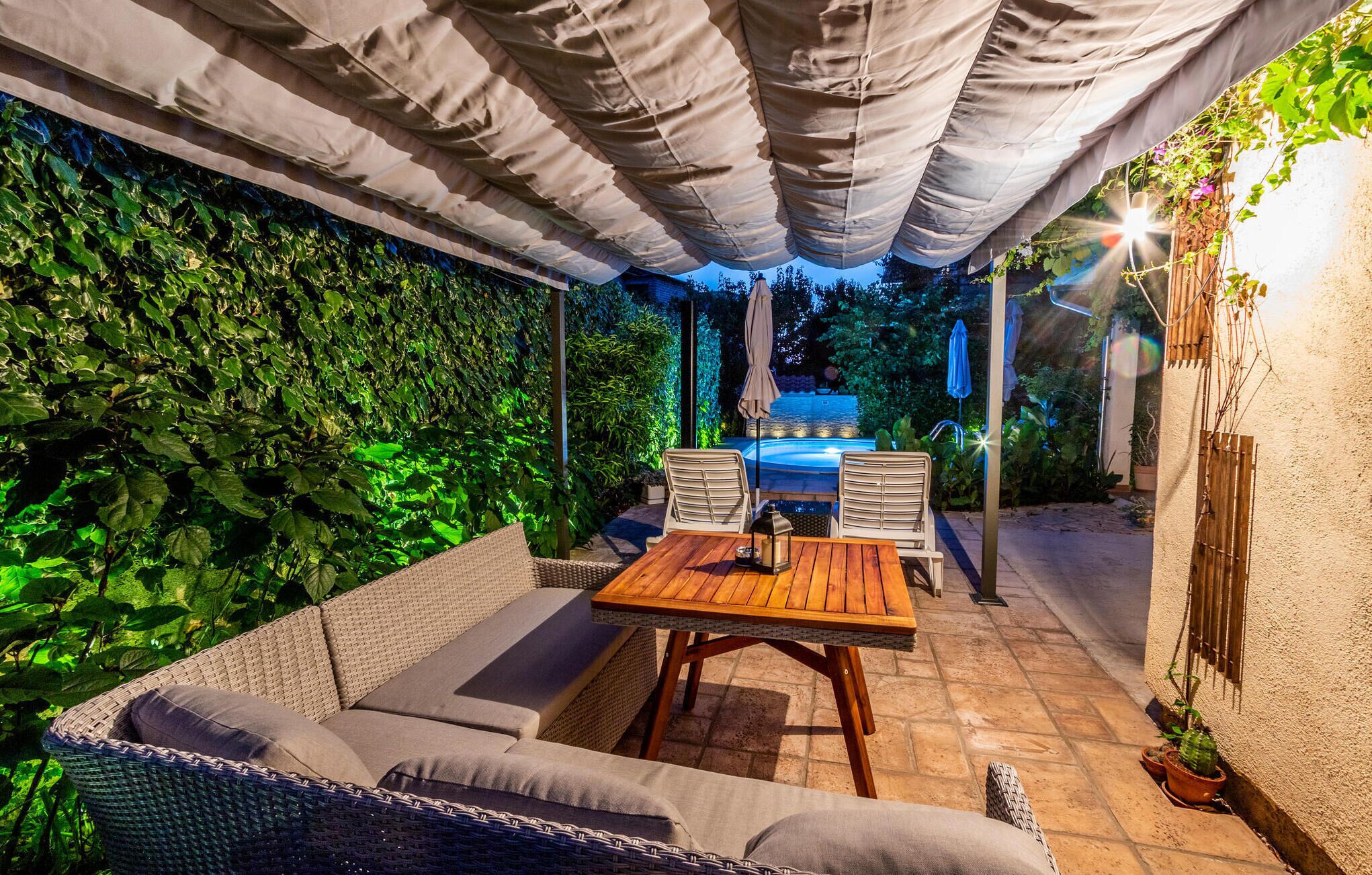 Eine Terrasse im Freien mit einem Holztisch und Korbmöbeln, umgeben von üppiger Bepflanzung und einem überdachten Bereich.