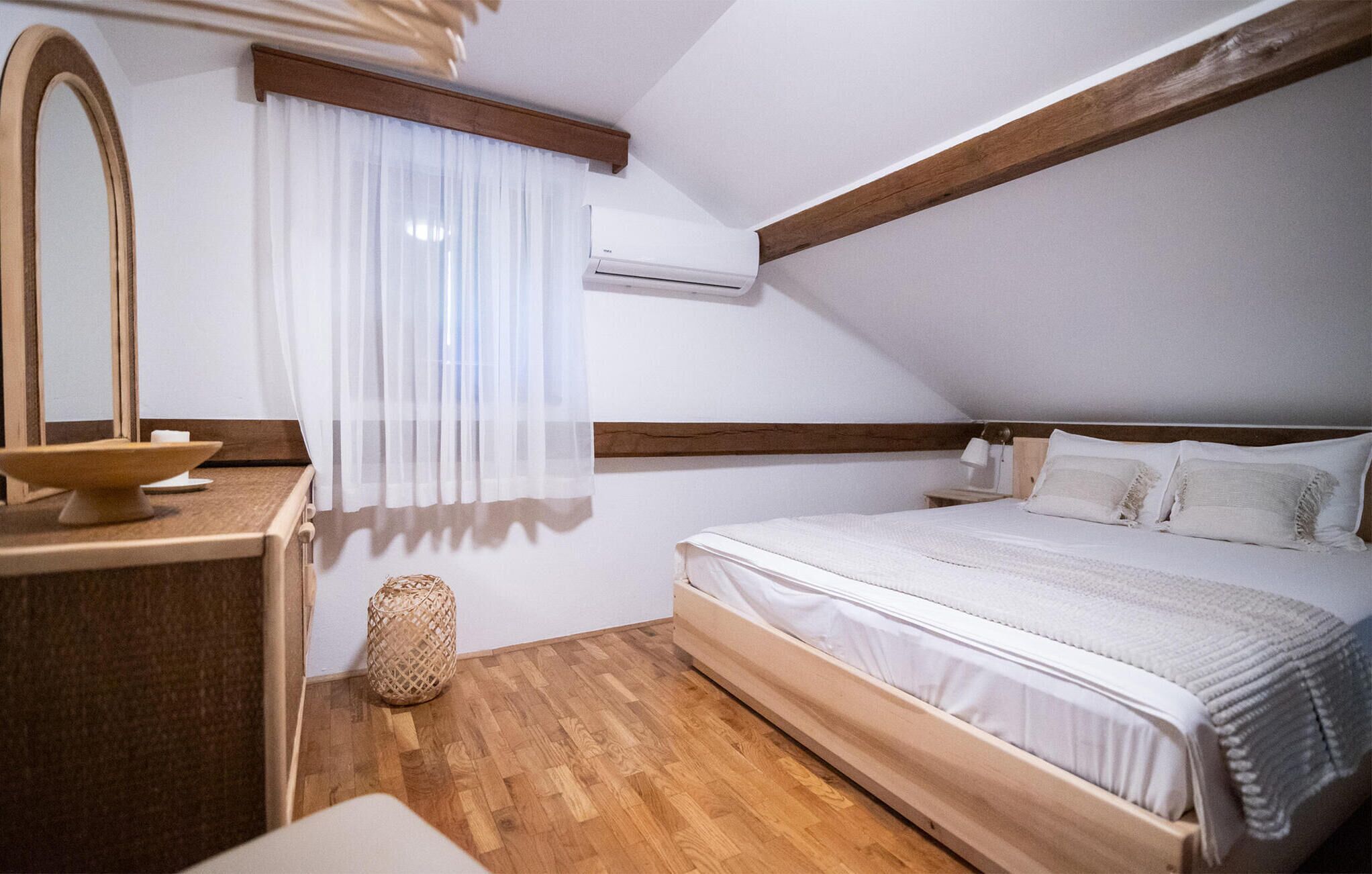 A bedroom with a wooden headboard, a bed with white bedding, a bedside table with a bowl, a mirror, and a window with sheer curtains.