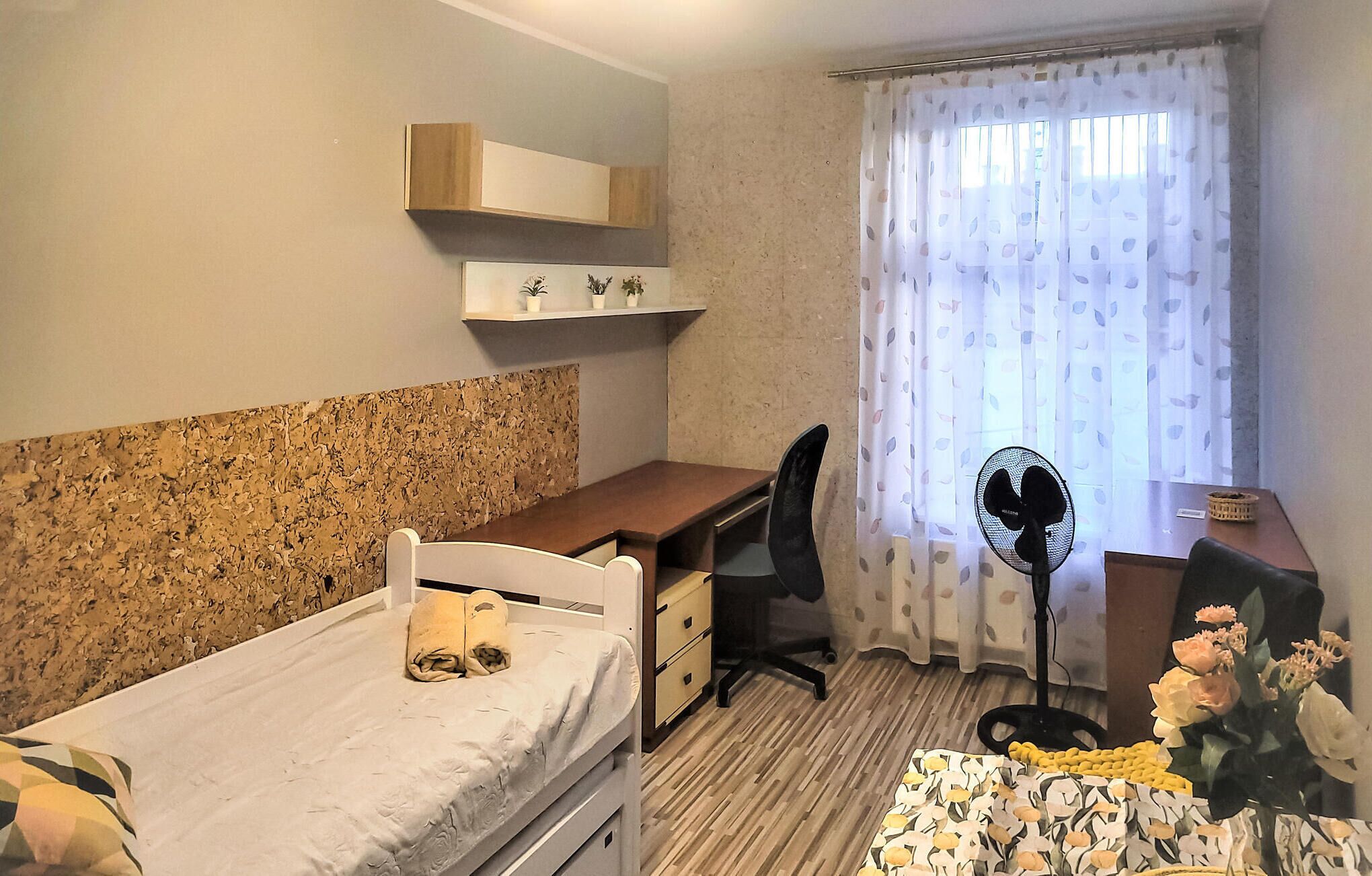 Ein kleiner, gut beleuchteter Raum mit einem Bett, einem Schreibtisch, einem Stuhl und einem Standventilator.