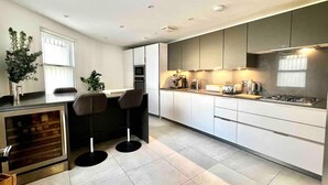 A modern kitchen with white cabinets, a built-in oven, and a gas stove. There are two bar stools at a small dining table, and a potted plant on the counter.