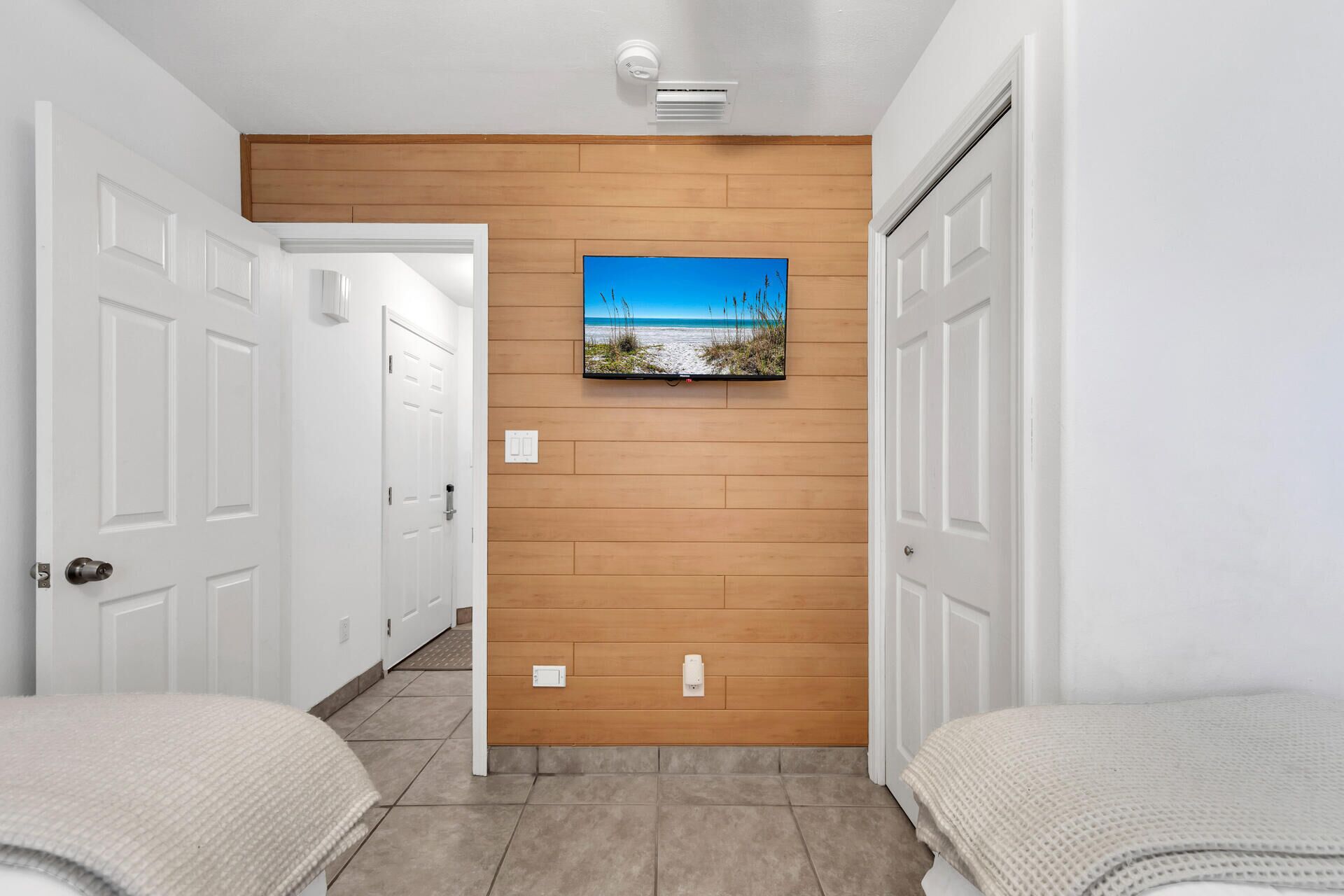 Un pasillo con una pared de acento de madera, un televisor montado que muestra una escena de playa y dos camas con ropa de cama color beige.